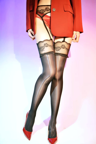 A woman posing in a black glossy thigh high stockings, with an attached garter belt with floral embroidery. 